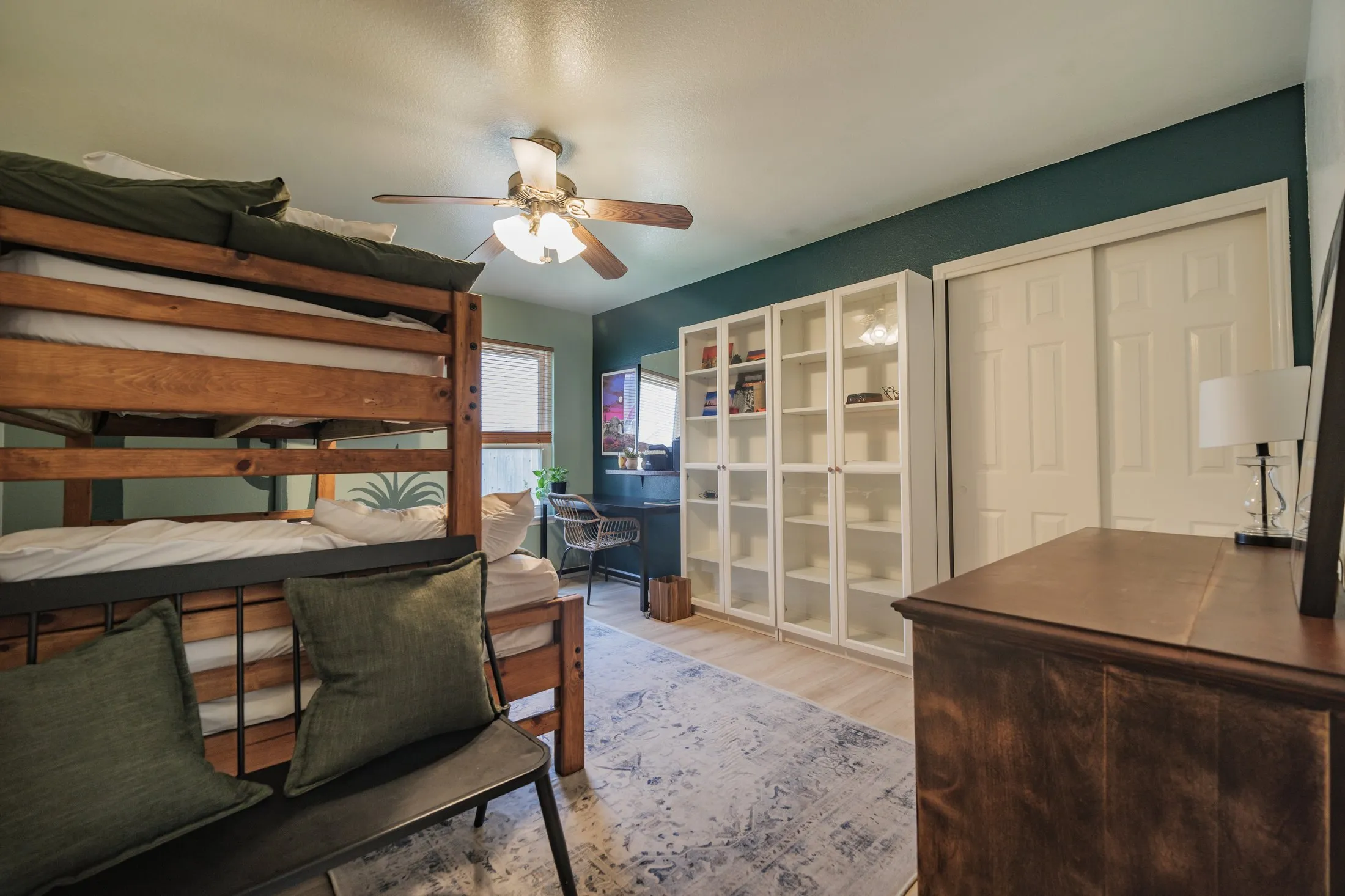 The bunk room: a solid wood bunk bed with green bedding, a deep teal accent wall, white glass-front bookcases, a desk with a chair, and a patterned rug on light wood flooring.