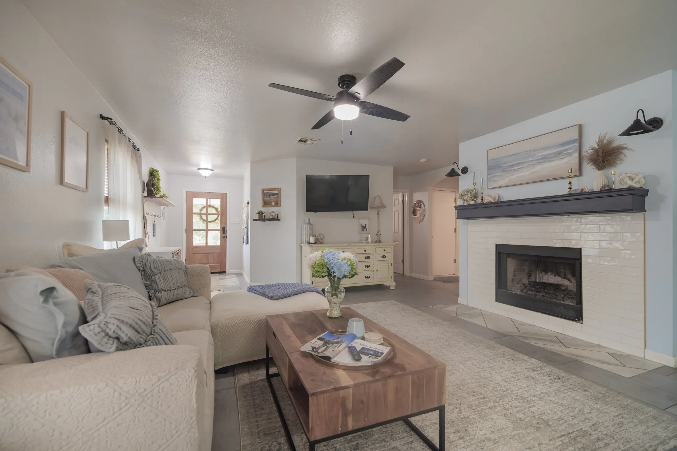 The living room: a cream sectional, pale blue accent wall, white subway-tile fireplace with a navy mantel, coastal landscape art, black barn-style sconces, and a wood coffee table on a soft grey rug.