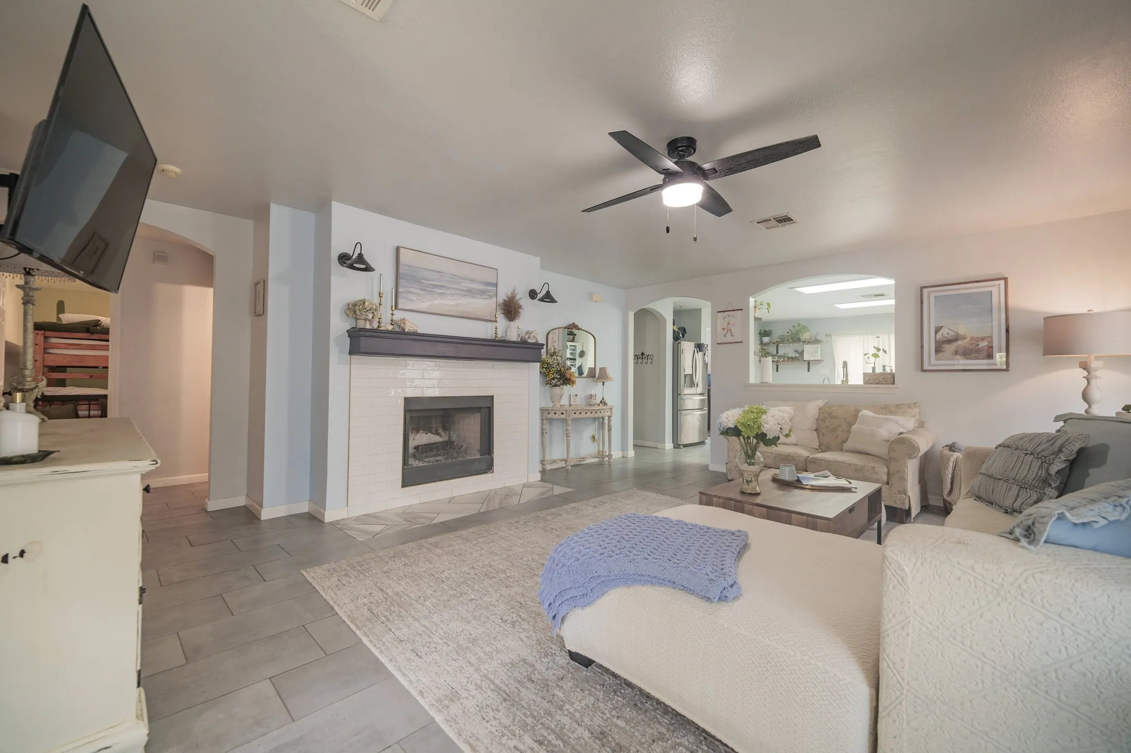 The open-plan living area after: pale blue walls, a white subway-tile fireplace with a navy mantel, coastal artwork, cream seating, and a glimpse through the archway into the lightened kitchen.