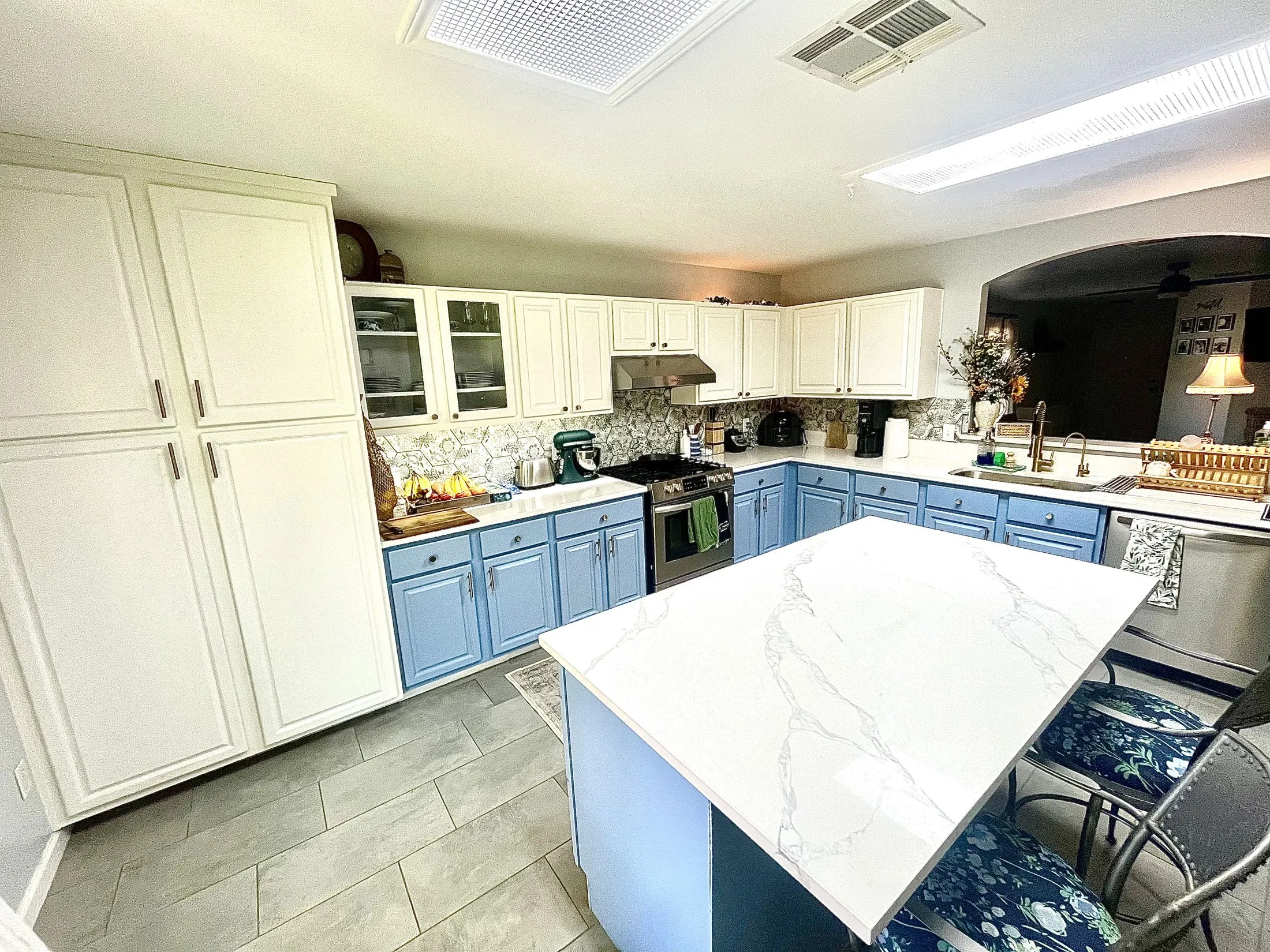 The kitchen before: blue painted lower cabinets, cream upper cabinets, a patterned hexagonal tile backsplash, fluorescent ceiling panels, and personal items across the counters.